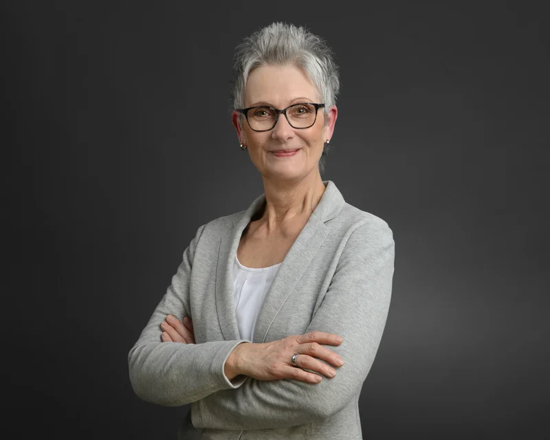 Portrait einer Frau mit kurzem grauem Haar und Brille im hellgrauen Blazer