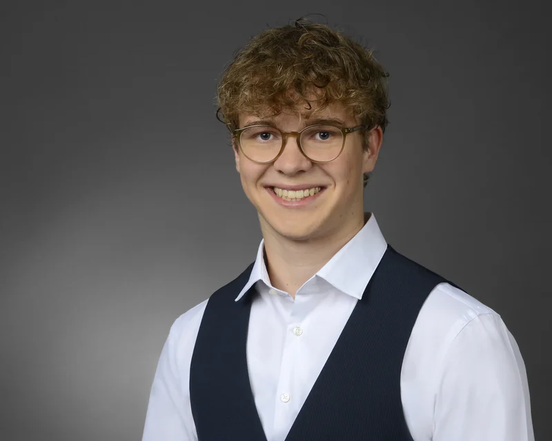 Bewerbungsportrait eines Mannes mit Locken und Brille in weißem Hemd und dunkler Weste